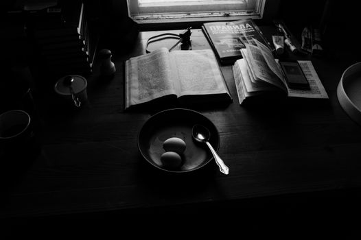 Black and white of plate with eggs and spoon near opened books and textbook and phone charger placed on wooden desk under light from window