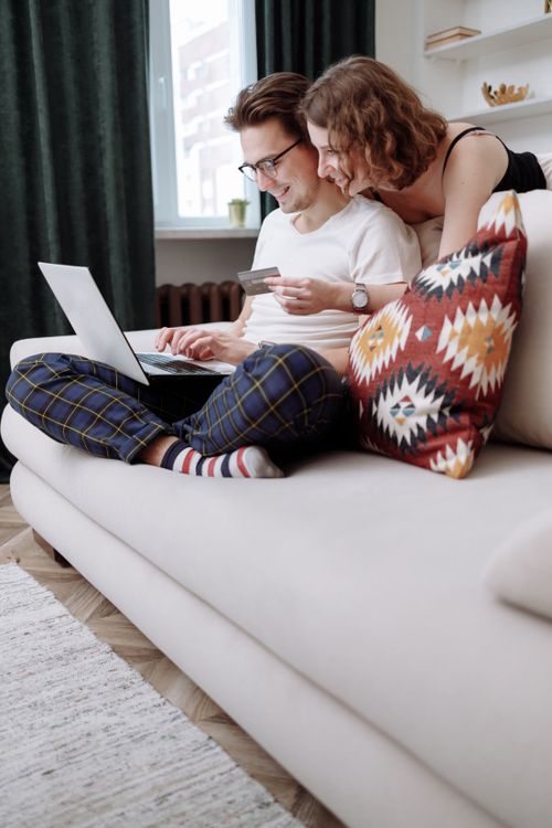 Free Happy couple sitting on a couch, using a laptop for online shopping with a credit card in hand. Stock Photo
