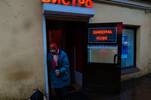 Senior gloomy female in shawl and warm jacket with cup of coffee leaning on entrance of bistro and looking at camera