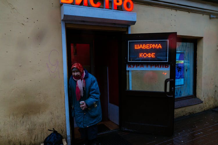 Aged Poor Distrustful Woman In Entrance Of Small Shop