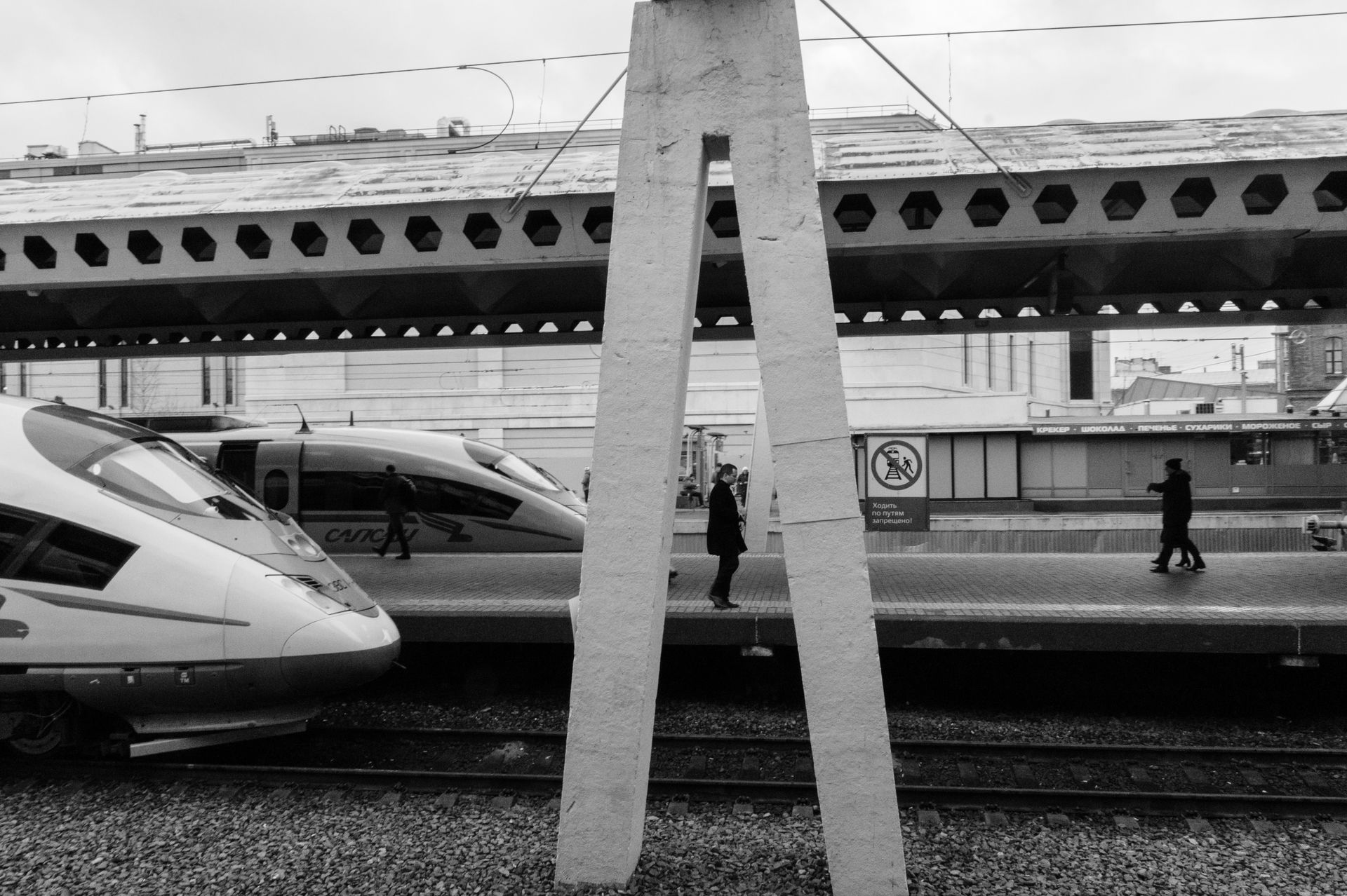 Black and white of station with speed locomotives on railroad and passengers walking along platform and stone support construction in center of image