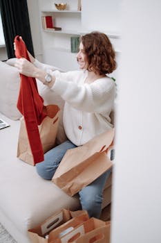 Happy woman sitting on a sofa, examining new clothes from shopping bags indoors.
