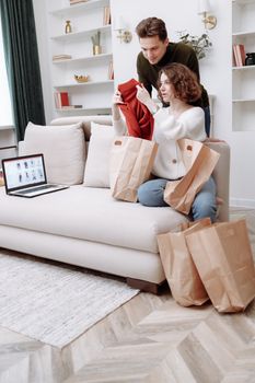 A couple enjoys online shopping in their living room surrounded by paper bags.