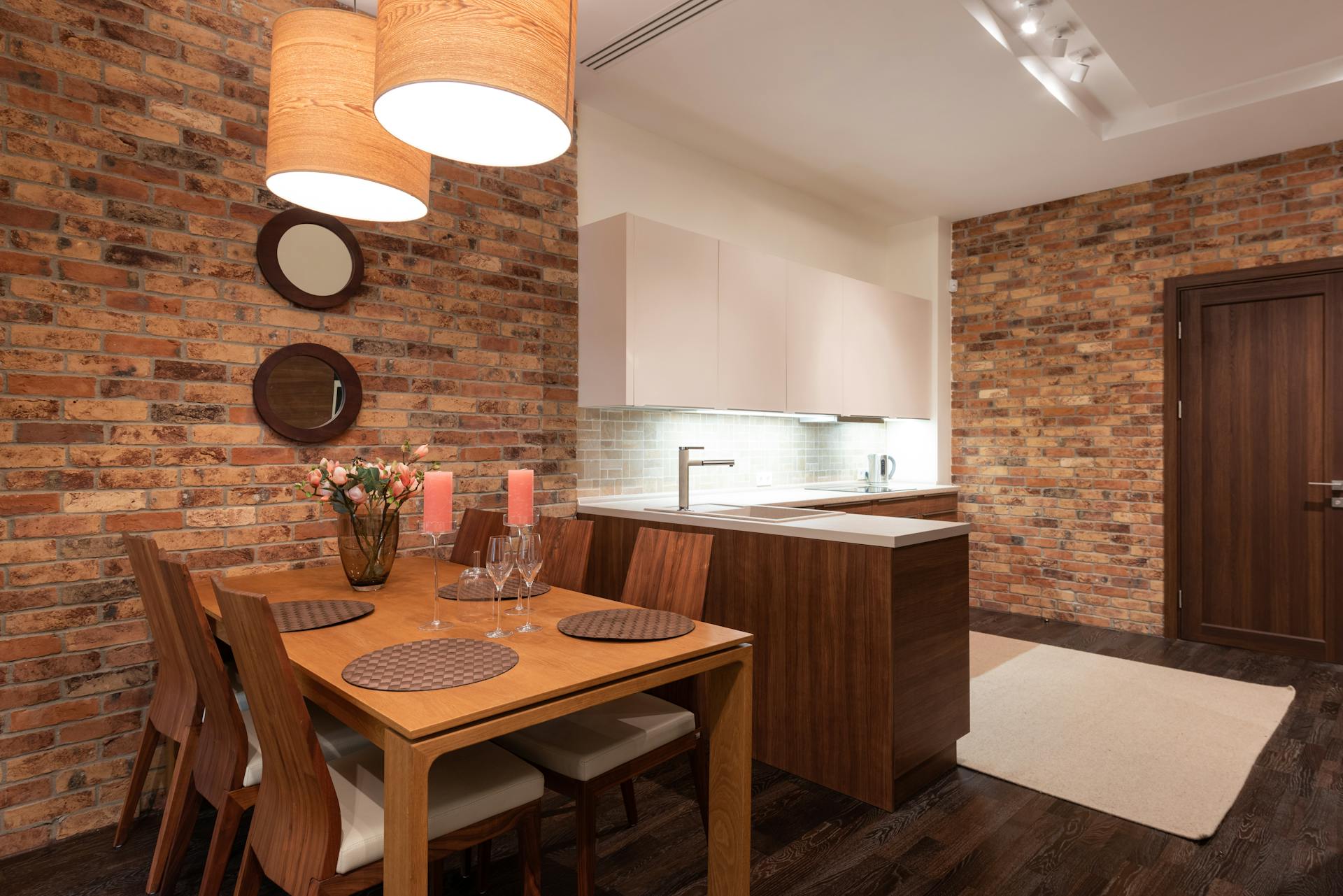 Light kitchen interior with table and chairs decorated with glasses and candles with flower vase and napkins near cabinets with tap and lamps on ceiling near brick walls and door