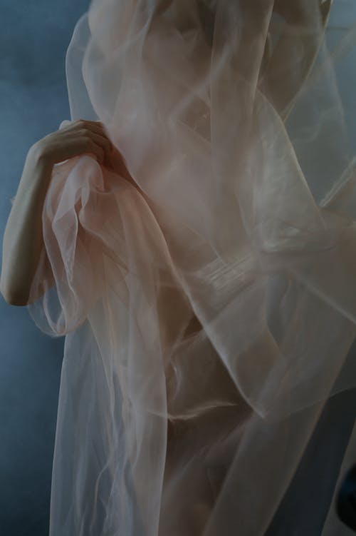 Crop anonymous female model covering naked body with translucent fabric and holding material with gentle hand