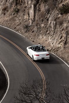 A white convertible car driving along a winding mountain road surrounded by rocky cliffs.