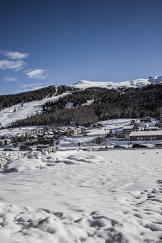 A picturesque snow-covered town in Livigno, Italy, with mountainous backdrop under a clear blue sky.