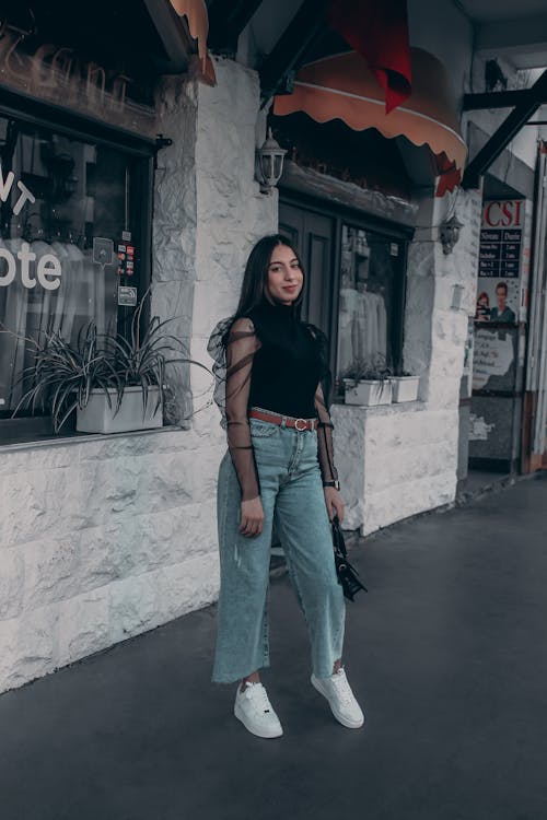 A Woman in Black Blouse and Denim Pants Standing Outside an Establishment while Smiling at the Camera