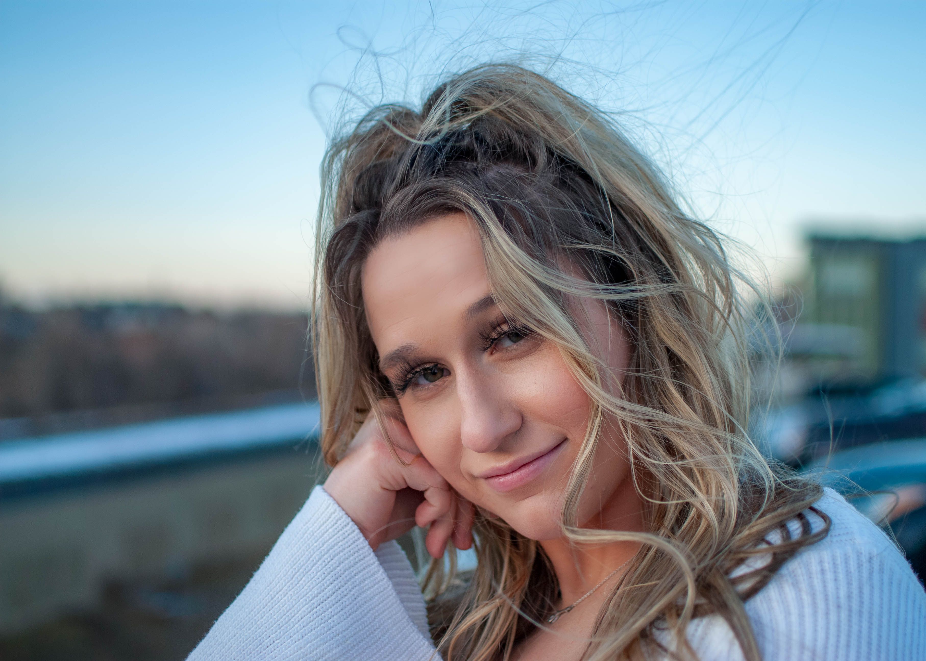 Close-up of a woman with blonde hair in a white sweater, smiling outdoors.