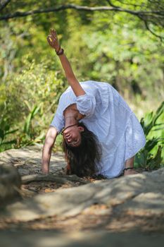 Full body of barefoot female in dress standing in Urdhva Dhanurasana posture with raised arm showing okay gesture in nature