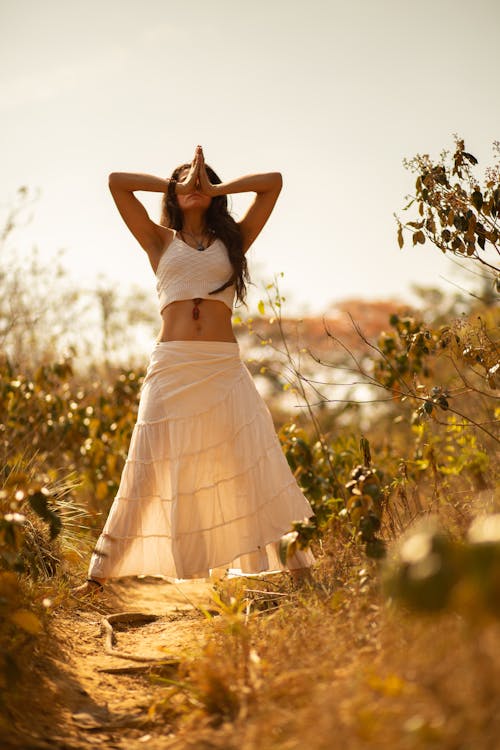 Full body of peaceful female in white summer outfit standing among plants with hands together during meditation