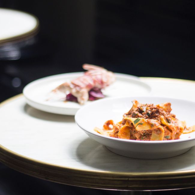 Round table with white bowl with delicious pasta with meat and herbs with sauce near plate with small roasted bird in light restaurant