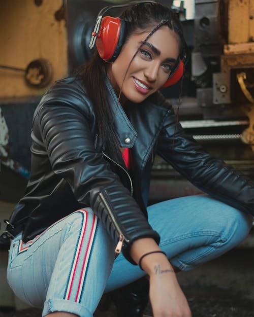 Woman in Black Leather Jacket and Blue Denim Jeans Wearing Red Headphones while Smiling at the Camera