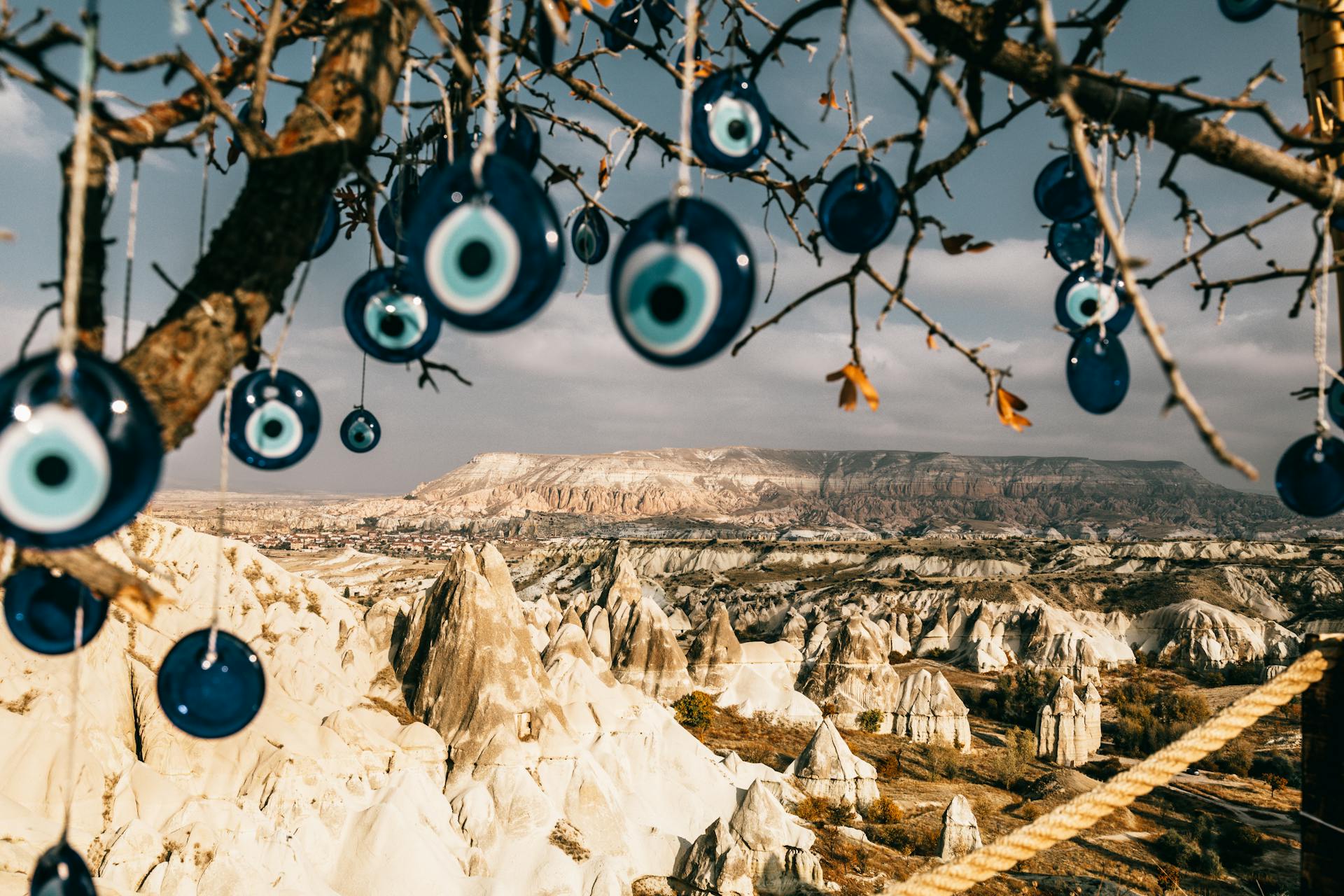 Nazar amulets on threads hanging from tree branches near rocky uneven formations with mountains and grass with plants in Turkey in Cappadocia region under gray cloudy sky in summer day