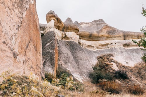 Uneven terrain of hills with stones surrounded with dry grass and green shrubs under cloudy gray sky