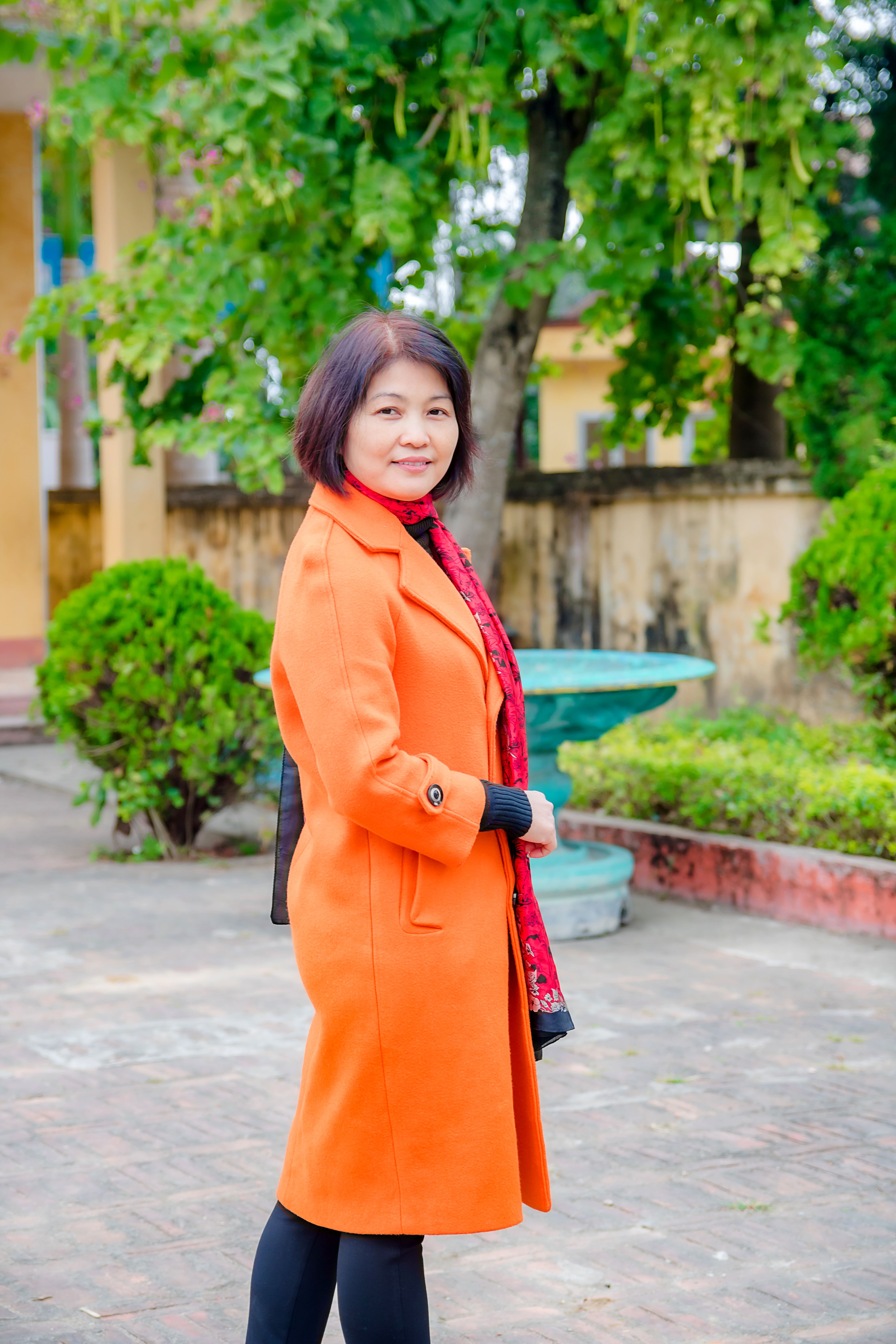 An elegant woman in orange coat poses in an outdoor garden setting with vibrant greenery.