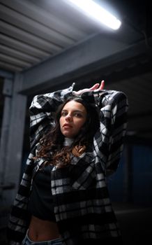Portrait of a young woman posing with arms raised in an industrial setting under a neon light.