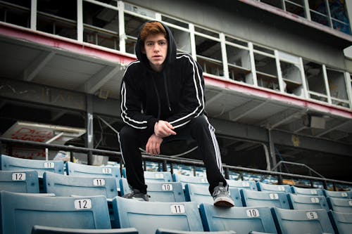 Photo of a Man in a Black Hoodie Crouching on Blue Bleachers