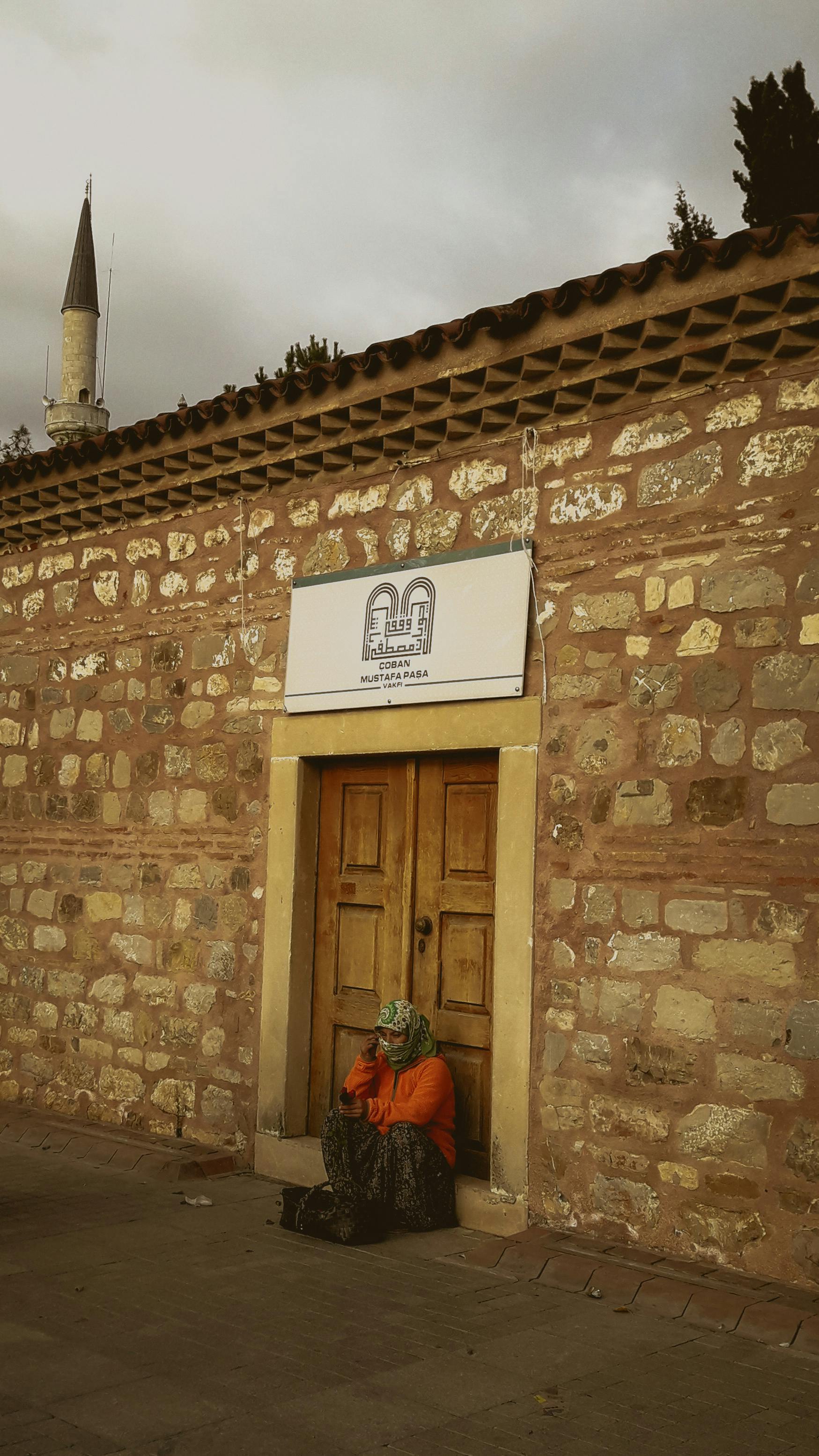 A woman in traditional attire sits by a rustic door against a stone wall, with a minaret in the background.