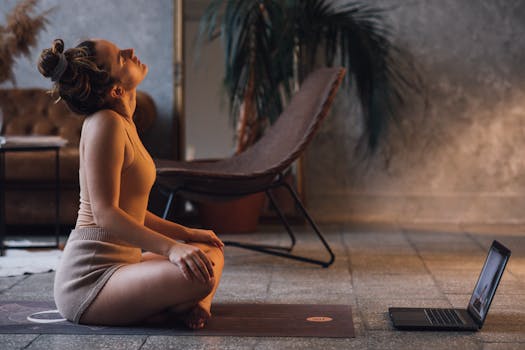 A woman doing yoga on a mat indoors, facing a laptop, in a serene setting.