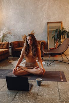 Woman meditating on a yoga mat with a laptop and mug in a stylish living room.