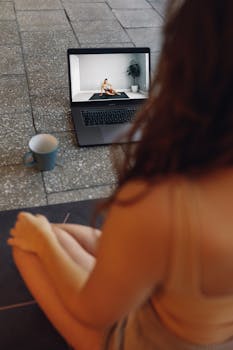 A woman follows a virtual yoga class at home, sitting on a mat with a laptop showing an instructor.