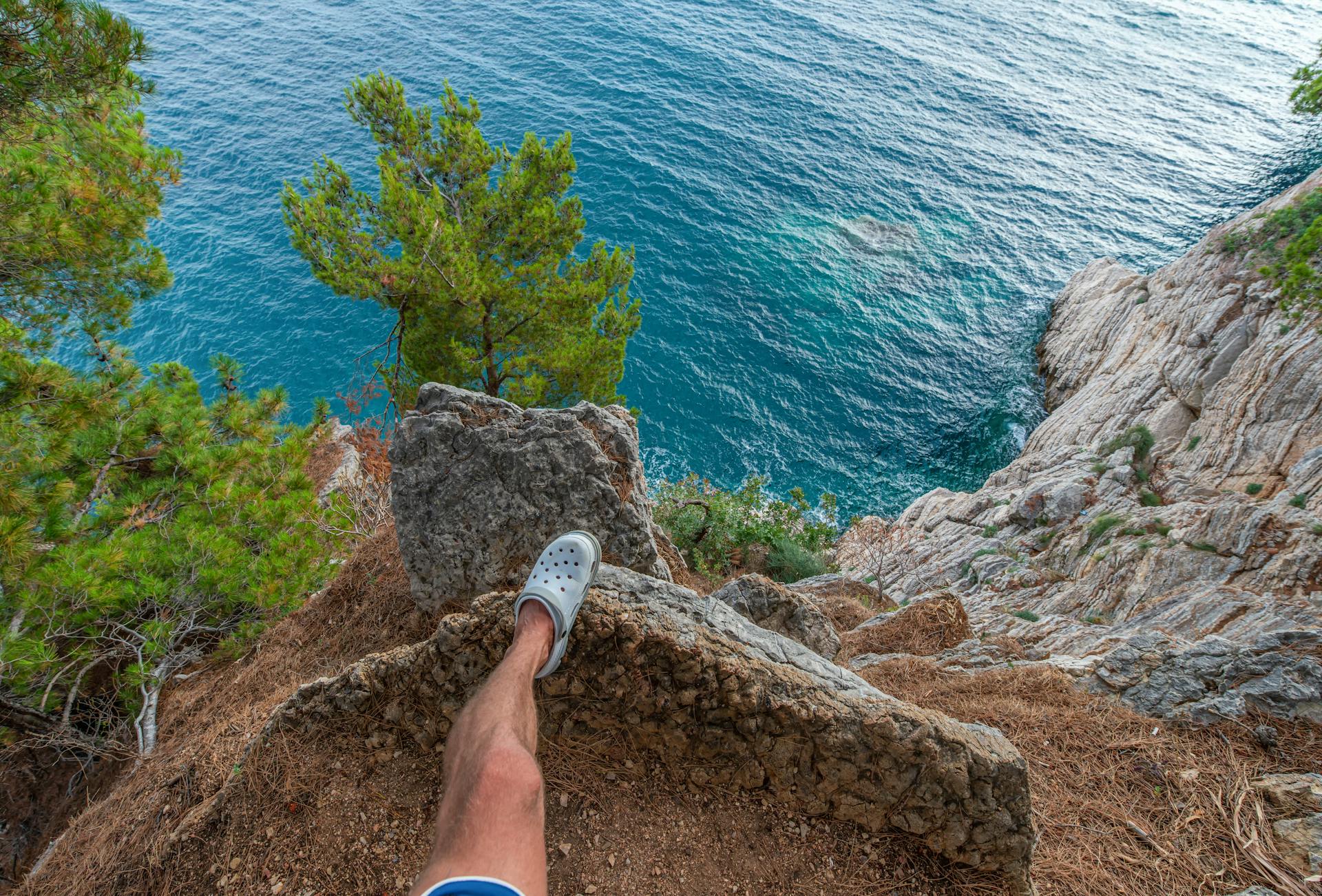 From above of anonymous male traveler standing on edge of rocky cliff above above azure rippling sea in tropical country