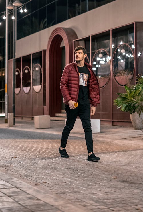 Man in Red Jacket and Black Pants Walking on the Side of the Road while Looking Afar