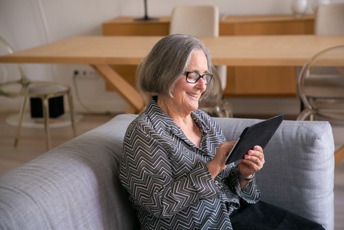 Free Elderly woman smiling while using a tablet in a cozy living room setting. Stock Photo