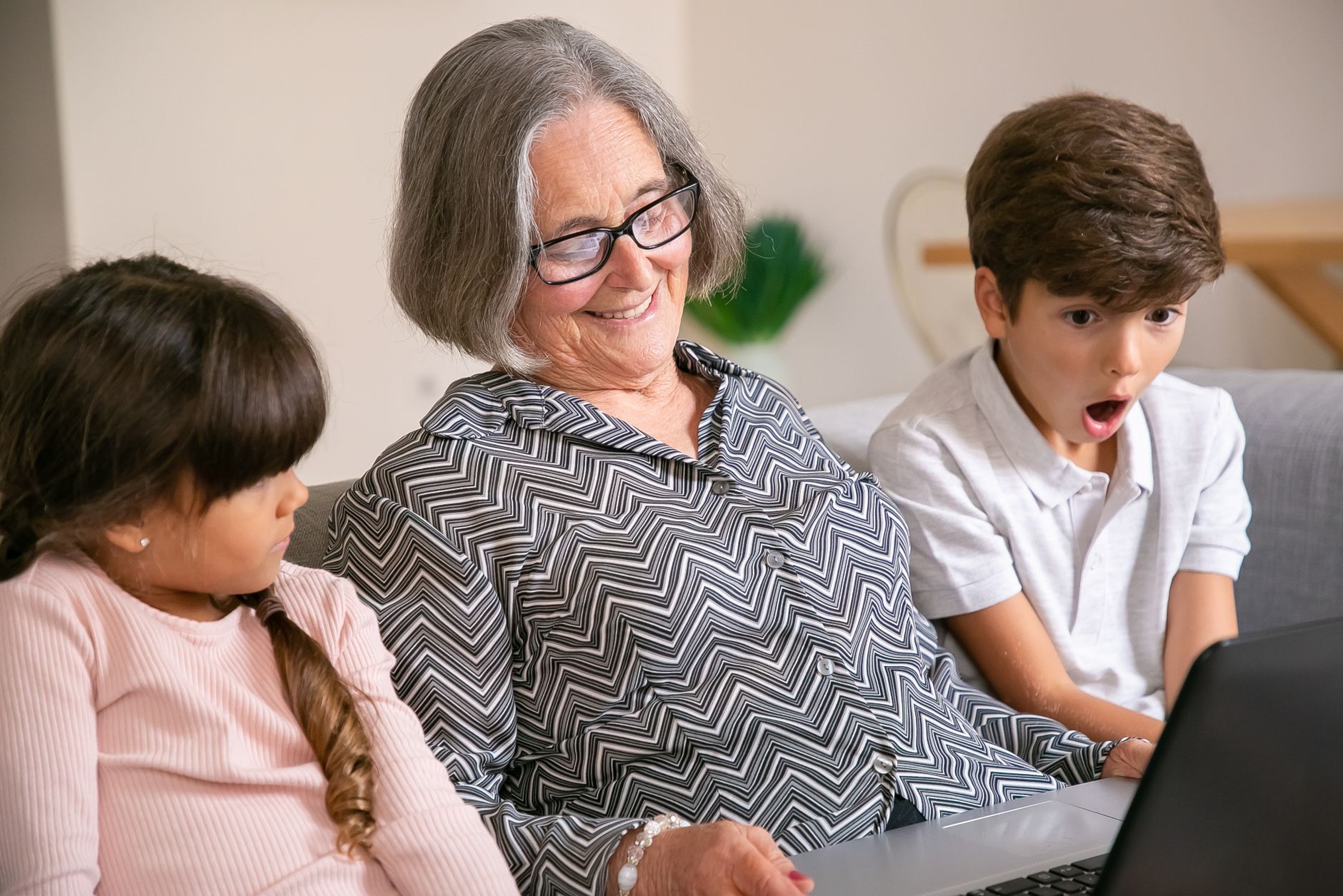 A grandmother with her grandchildren joyfully looking at a laptop indoors.