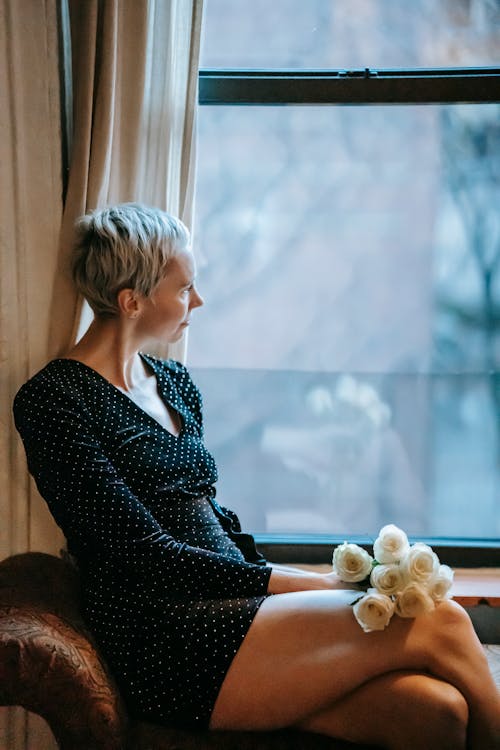 Free Side view of thoughtful woman with short hair in black dress sitting with bouquet of white roses near window and looking away Stock Photo