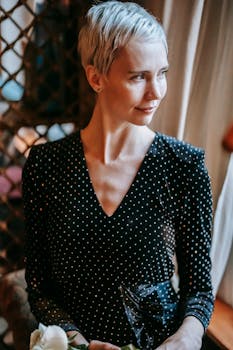 Positive woman with short blond hairstyle in stylish outfit standing with fresh white rose in hands and looking away in room in daytime
