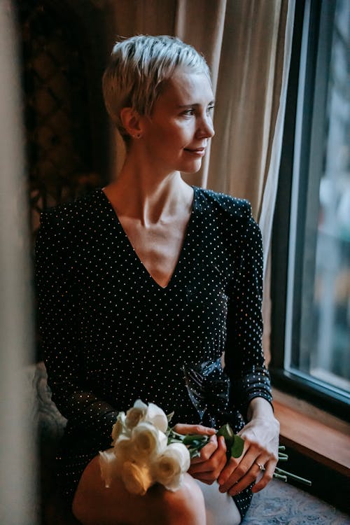 Free Feminine adult lady with short dyed hair in elegant dress resting on windowsill with bouquet of roses and looking away Stock Photo