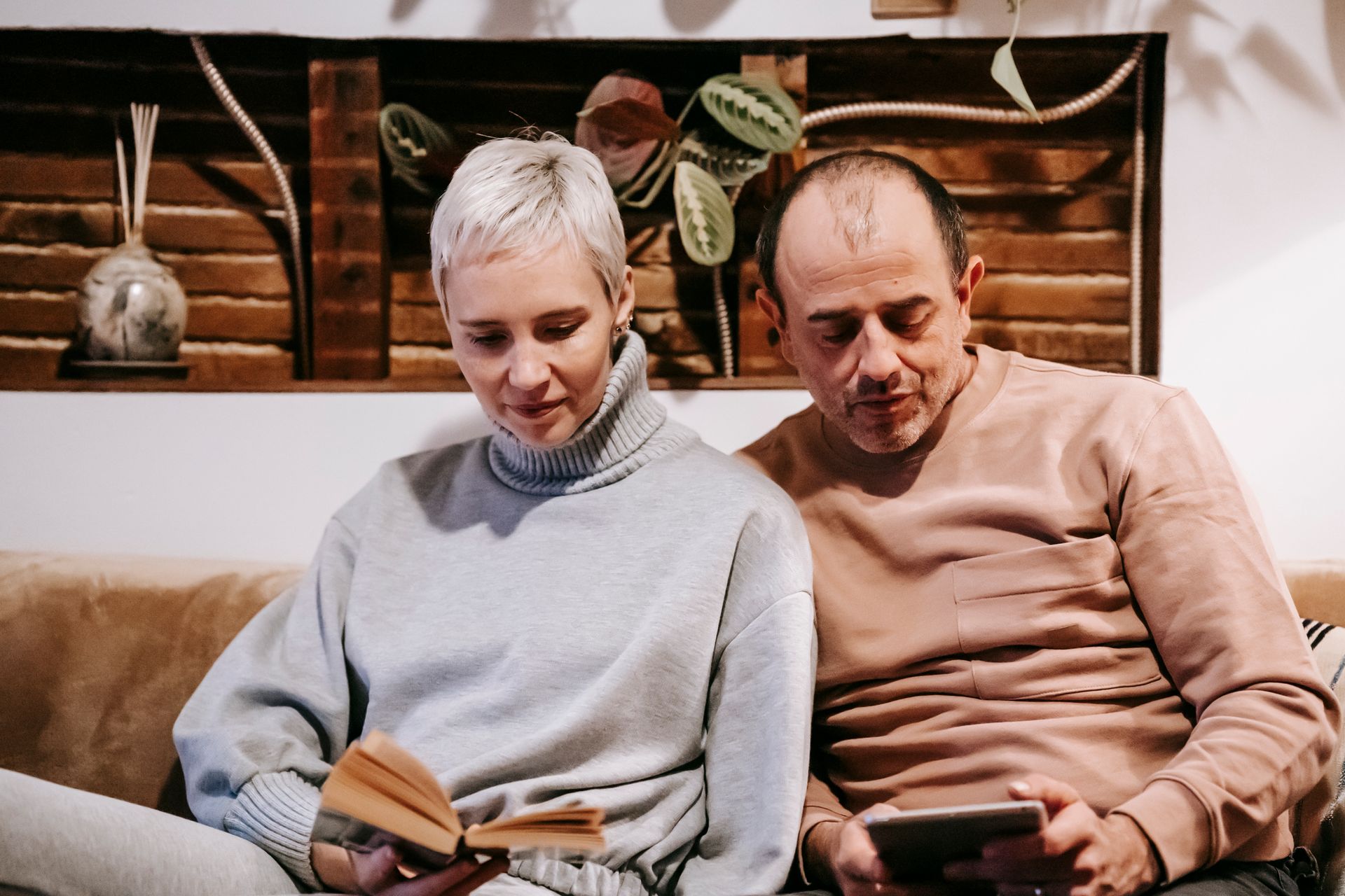 Concentrated middle aged diverse spouses in casual clothes sitting together on sofa while reading book and using tablet during weekend at home