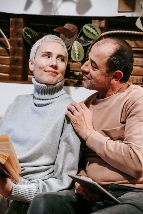 Free Positive diverse middle aged spouses in warm sweaters smiling and looking at each other while resting on sofa with book and tablet Stock Photo