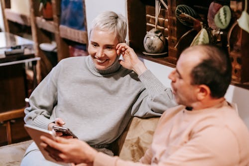 From above of middle aged diverse in casual clothes smiling while sitting on sofa and using smartphone and tablet during weekend at home