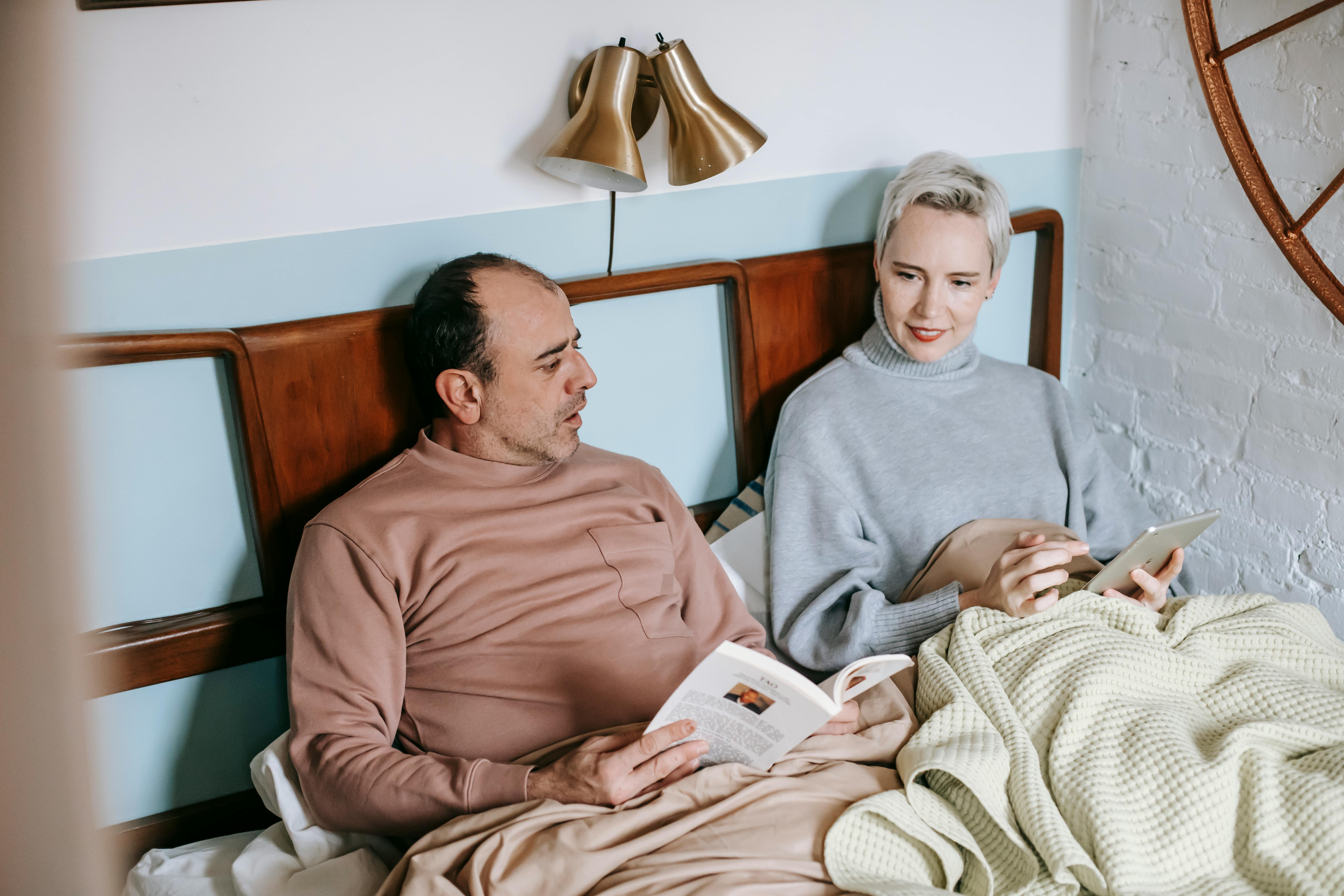 adult woman using table while sitting on bed near ethnic husband reading novel