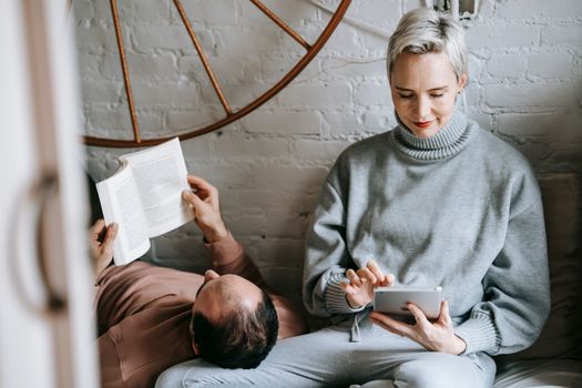 Anonymous man in casual clothes reading book while lying on knees of happy girlfriend using tablet at home