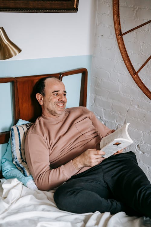 Free Happy middle aged ethnic male in casual clothes smiling while relaxing on bed and reading interesting book Stock Photo