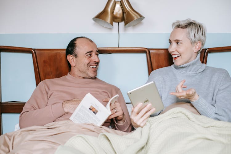 Cheerful Diverse Couple Relaxing In Bed With Book And Tablet During Lazy Weekend