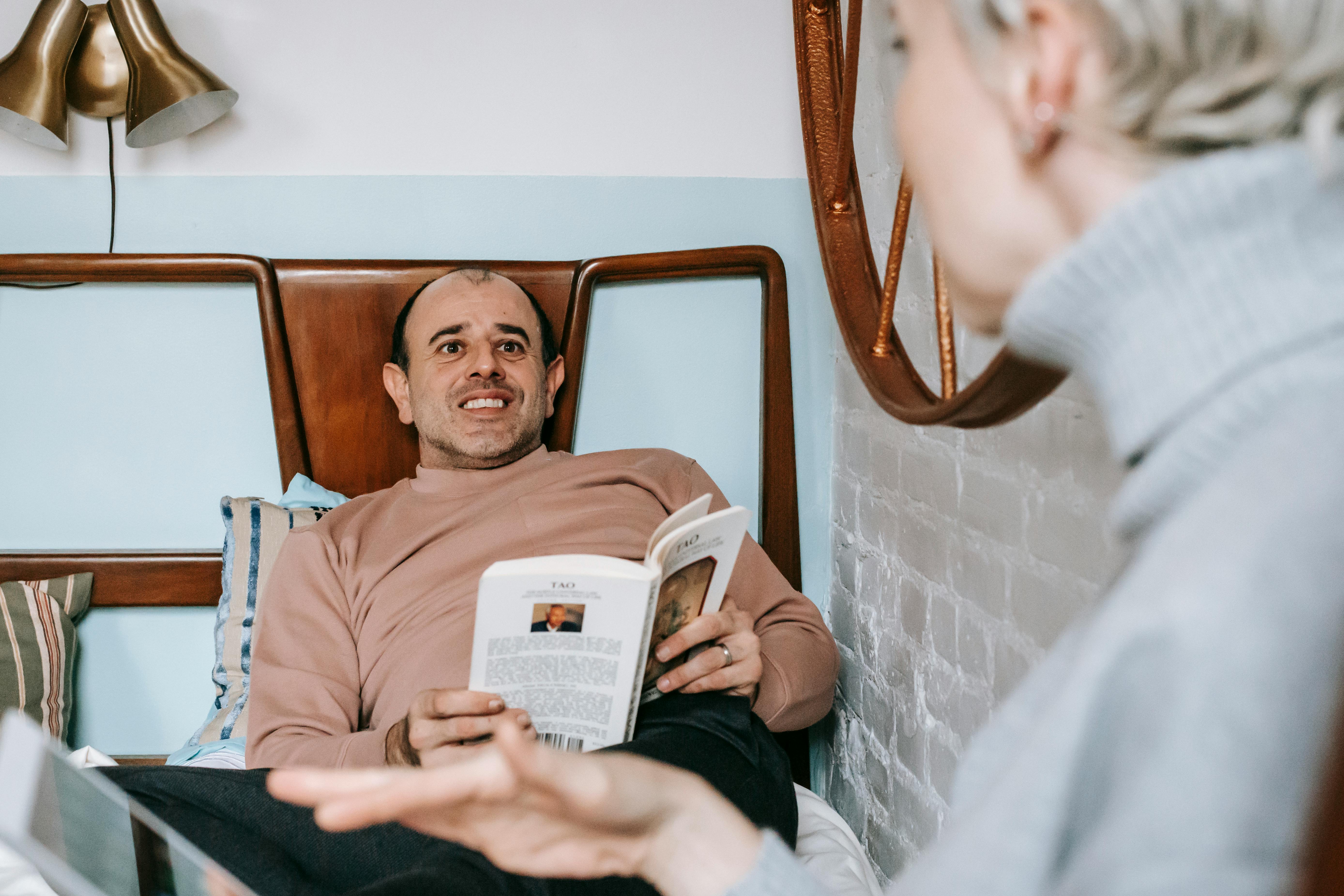 smiling ethnic man reading book on bed near wife surfing tablet