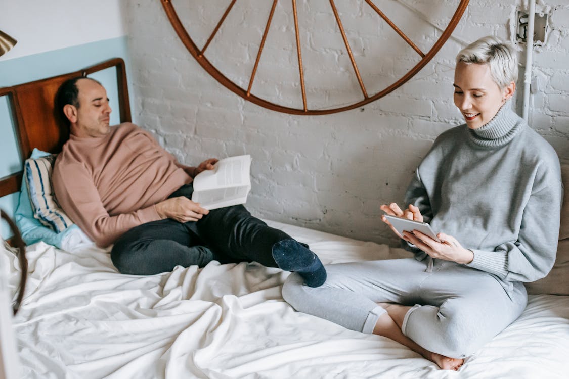 Positive woman surfing tablet on bed near husband reading book