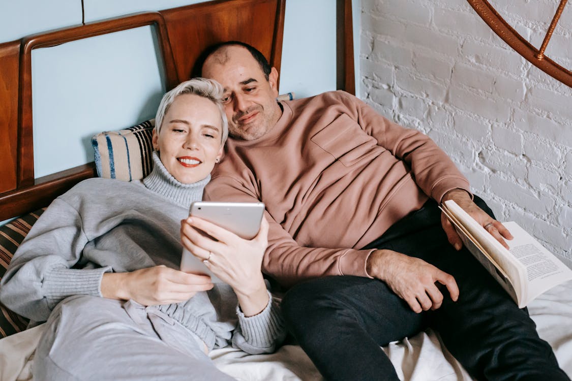 Free From above of happy adult diverse couple in casual outfits lying on bed and sharing table during weekend at home Stock Photo