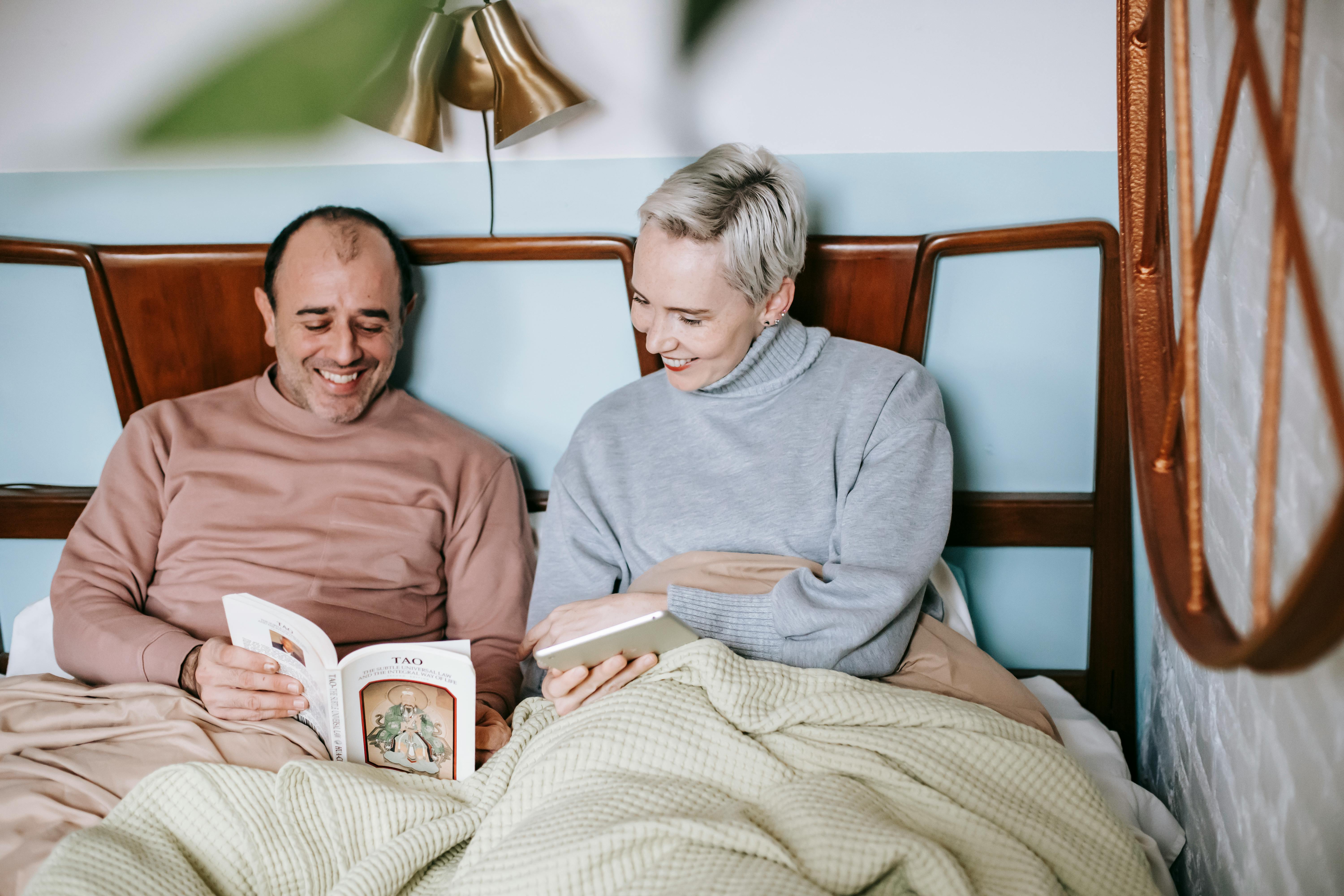 happy diverse couple smiling and reading book together while resting in bed