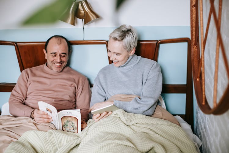 Happy Diverse Couple Smiling And Reading Book Together While Resting In Bed