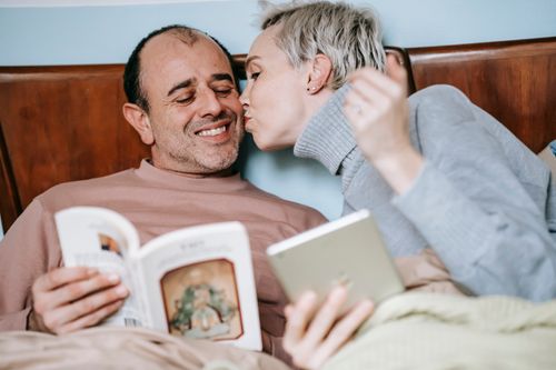 Free Loving adult woman in casual clothes with tablet in hand kissing cheek of smiling ethic husband reading book in bed Stock Photo