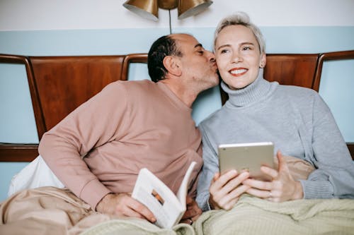 Free Romantic ethnic man kissing cheek of happy wife while recreating in bed Stock Photo