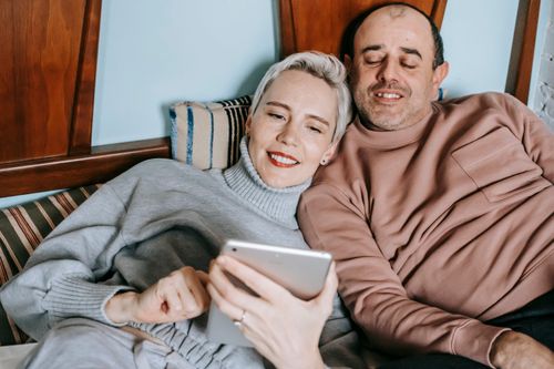 Free Content diverse middle aged married couple in warm casual clothes lying together on bed and smiling while watching movie on tablet Stock Photo