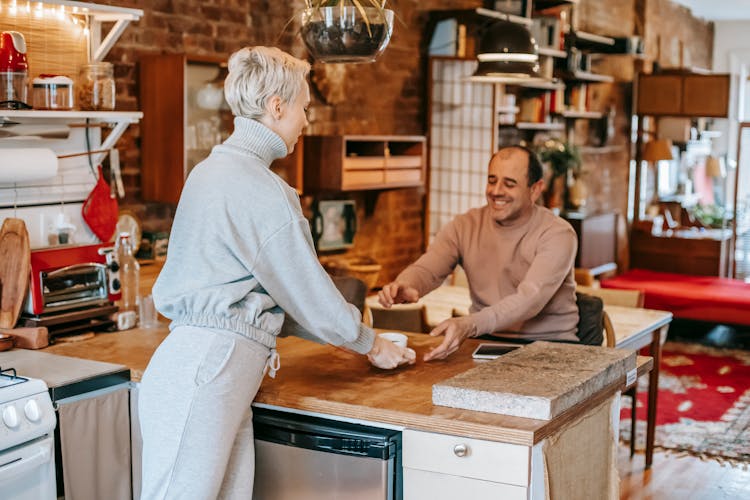 Woman Passing Coffee To Smiling Friend In House