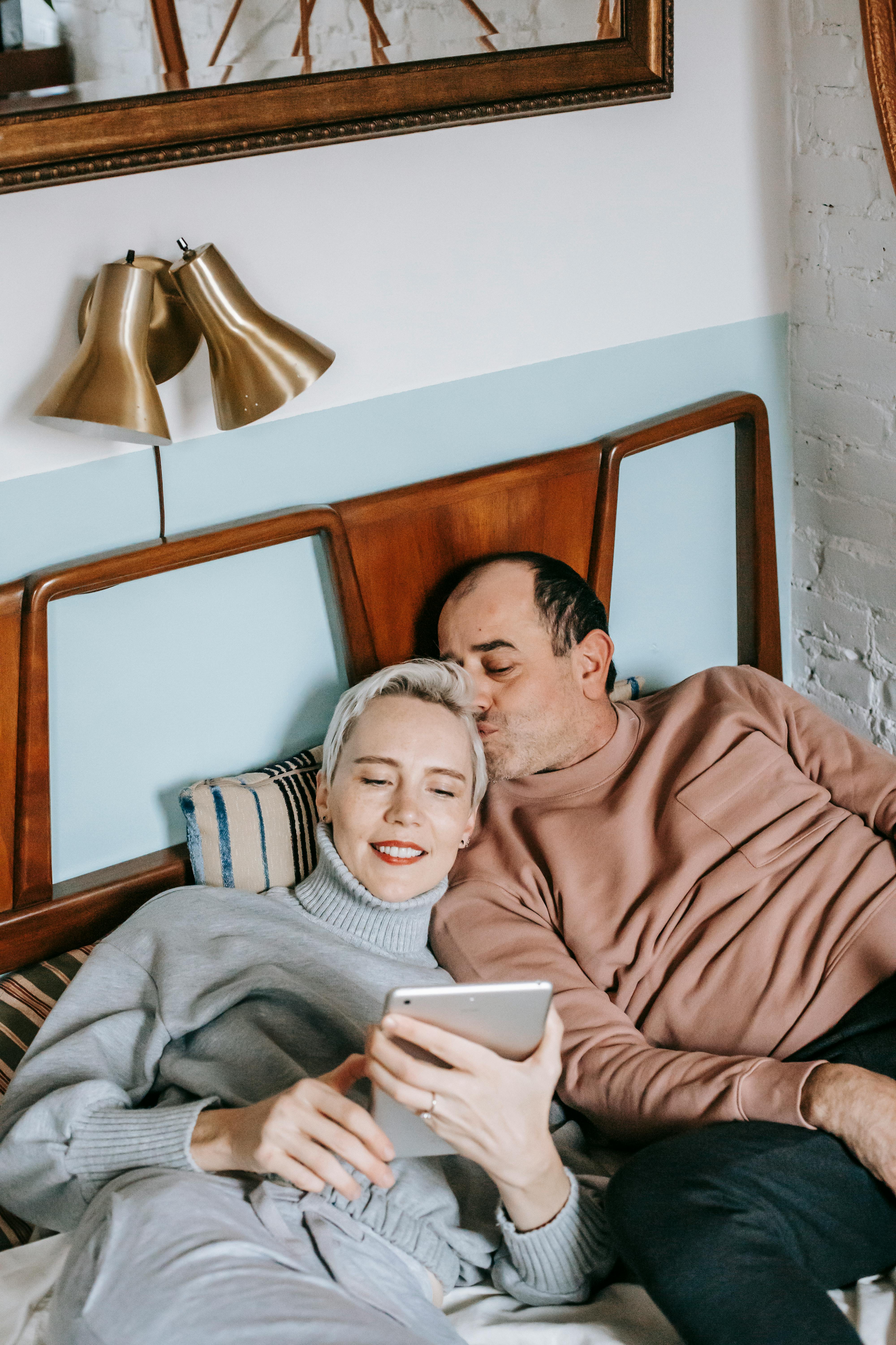 happy ethnic man kissing head of wife using tablet on bed